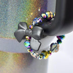 A black bow-shaped tumbler handle charm is attached to a black object, with a beaded bracelet wrapped around it. The bracelet features iridescent, faceted beads in shades of purple, blue, and yellow. The background has a glittery, multicolored effect.
