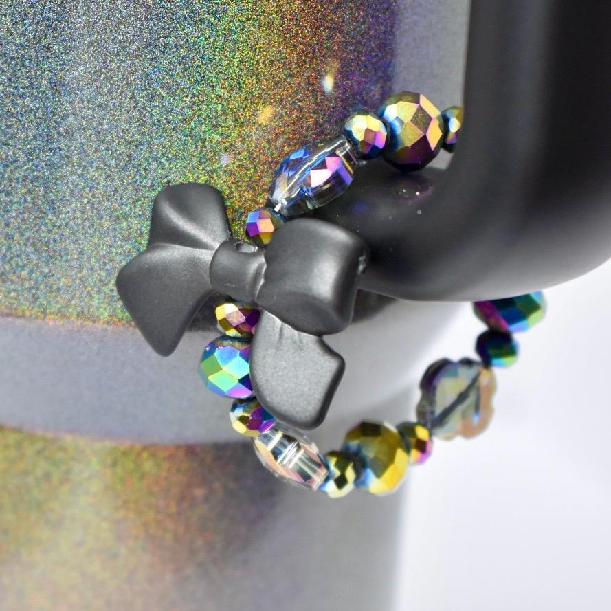 A black bow-shaped tumbler handle charm is attached to a black object, with a beaded bracelet wrapped around it. The bracelet features iridescent, faceted beads in shades of purple, blue, and yellow. The background has a glittery, multicolored effect.
