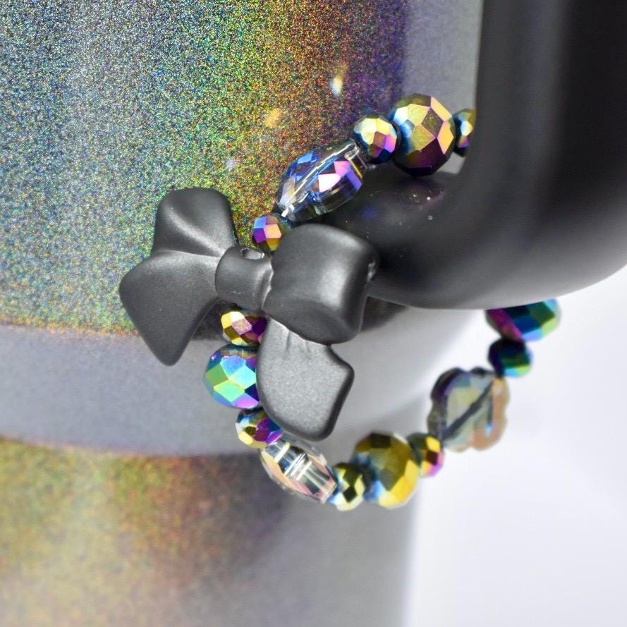 A black bow-shaped tumbler handle charm is attached to a black object, with a beaded bracelet wrapped around it. The bracelet features iridescent, faceted beads in shades of purple, blue, and yellow. The background has a glittery, multicolored effect.