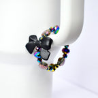 A black bow tumbler handle bracelet with iridescent beads. The bow is made of a matte black material and is the focal point of the bracelet. The beads are multi-colored, reflecting hues of purple, green, and gold. The bracelet is displayed on a white Stanley tumbler.