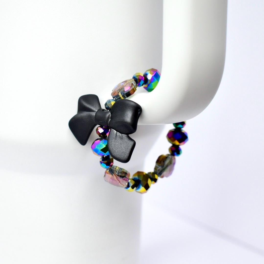 A black bow tumbler handle bracelet with iridescent beads. The bow is made of a matte black material and is the focal point of the bracelet. The beads are multi-colored, reflecting hues of purple, green, and gold. The bracelet is displayed on a white Stanley tumbler.