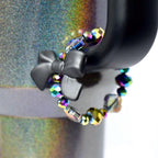 A black bow-shaped tumbler handle charm is attached to a dark, iridescent tumbler. A beaded bracelet wraps around the tumbler, featuring iridescent, multi-faceted beads in shades of purple, green, and yellow. The tumbler has a black handle.