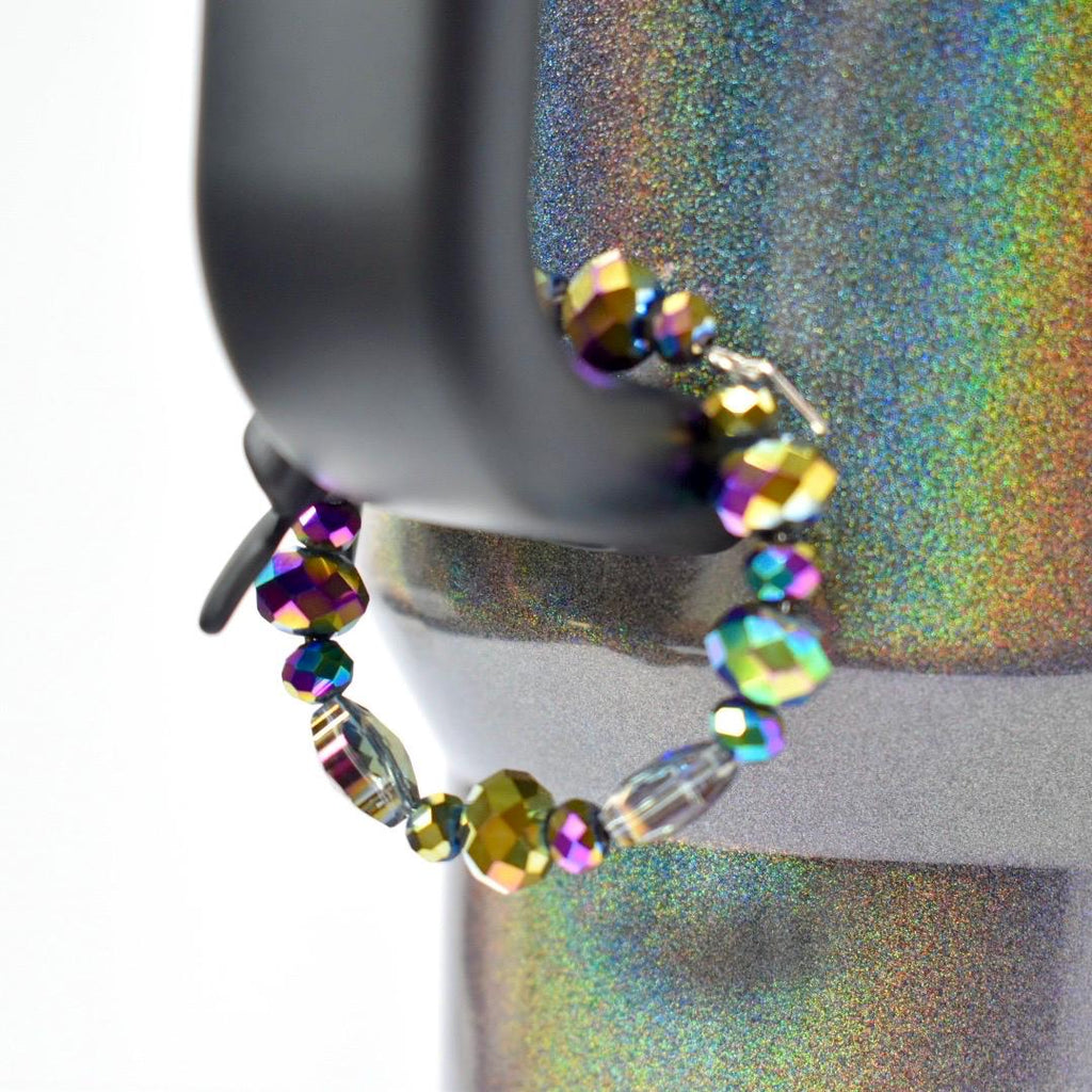 A beaded bracelet with iridescent beads. The bracelet is strung on a silver-colored wire and features a variety of bead shapes, including round and faceted. The beads display a range of colors, including purple, green, and gold, creating a shimmering effect. The bracelet is displayed on a black object.