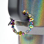 A beaded bracelet with iridescent beads. The bracelet is strung on a silver-colored wire and features a variety of bead shapes, including round and faceted. The beads display a range of colors, including purple, green, and gold, creating a shimmering effect. The bracelet is displayed on a black object.