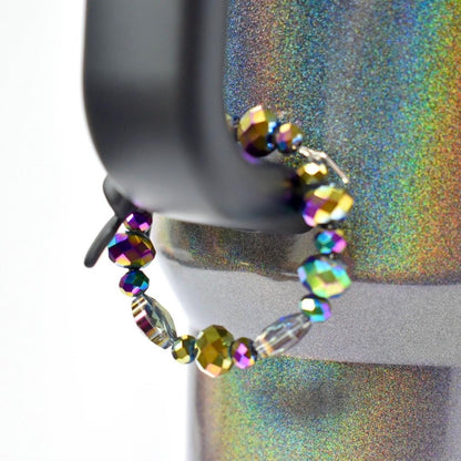 A beaded bracelet with iridescent beads. The bracelet is strung on a silver-colored wire and features a variety of bead shapes, including round and faceted. The beads display a range of colors, including purple, green, and gold, creating a shimmering effect. The bracelet is displayed on a black object.