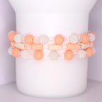 Three stackable tumbler bracelets featuring a combination of peach matte beads, clear faceted beads, white textured pearl-like beads, and cream-colored disc beads, arranged in a repeating pattern around a white tumbler.