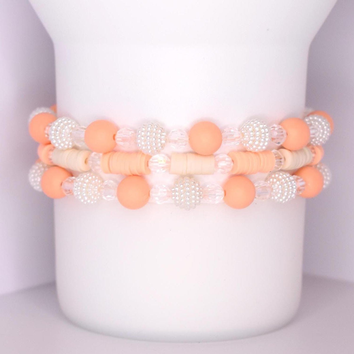 Three stackable tumbler bracelets featuring a combination of peach matte beads, clear faceted beads, white textured pearl-like beads, and cream-colored disc beads, arranged in a repeating pattern around a white tumbler.
