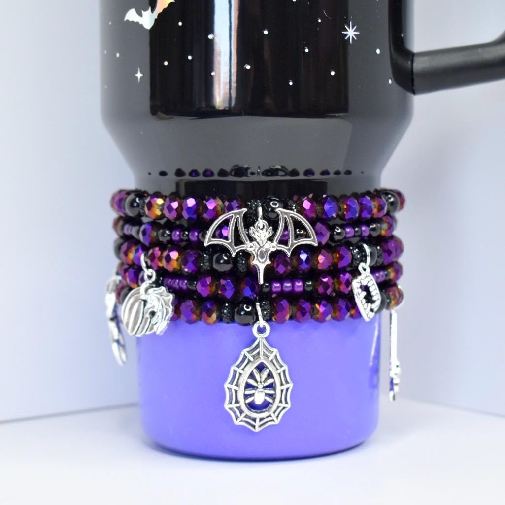 A black and purple tumbler with a beaded wrap. The wrap features purple and black beads, silver charms of a bat, spiderweb, pumpkin, and other Halloween-themed charms. The tumbler has a black top with a bat and stars.