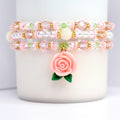 Three beaded bracelets in pink, white, and gold. Each bracelet features a mix of beads, including clear, pink, and white beads, with gold accents. A pink rose charm with green leaves hangs from the bottom bracelet. The bracelets are stacked on a white surface.
