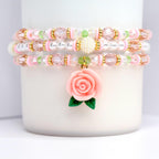 Three beaded bracelets in pink, white, and gold. Each bracelet features a mix of beads, including clear, pink, and white beads, with gold accents. A pink rose charm with green leaves hangs from the bottom bracelet. The bracelets are stacked on a white surface.