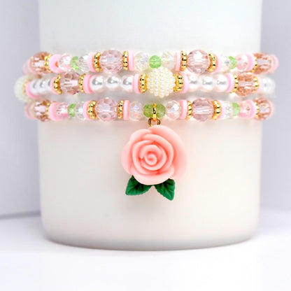 Three beaded bracelets in pink, white, and gold. Each bracelet features a mix of beads, including clear, pink, and white beads, with gold accents. A pink rose charm with green leaves hangs from the bottom bracelet. The bracelets are stacked on a white surface.