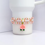 A beaded bracelet set with three strands. The bracelets are pink, white, and gold, with clear and pink beads. A pink rose charm with green leaves hangs from the bottom strand. The bracelets are displayed on a white surface.