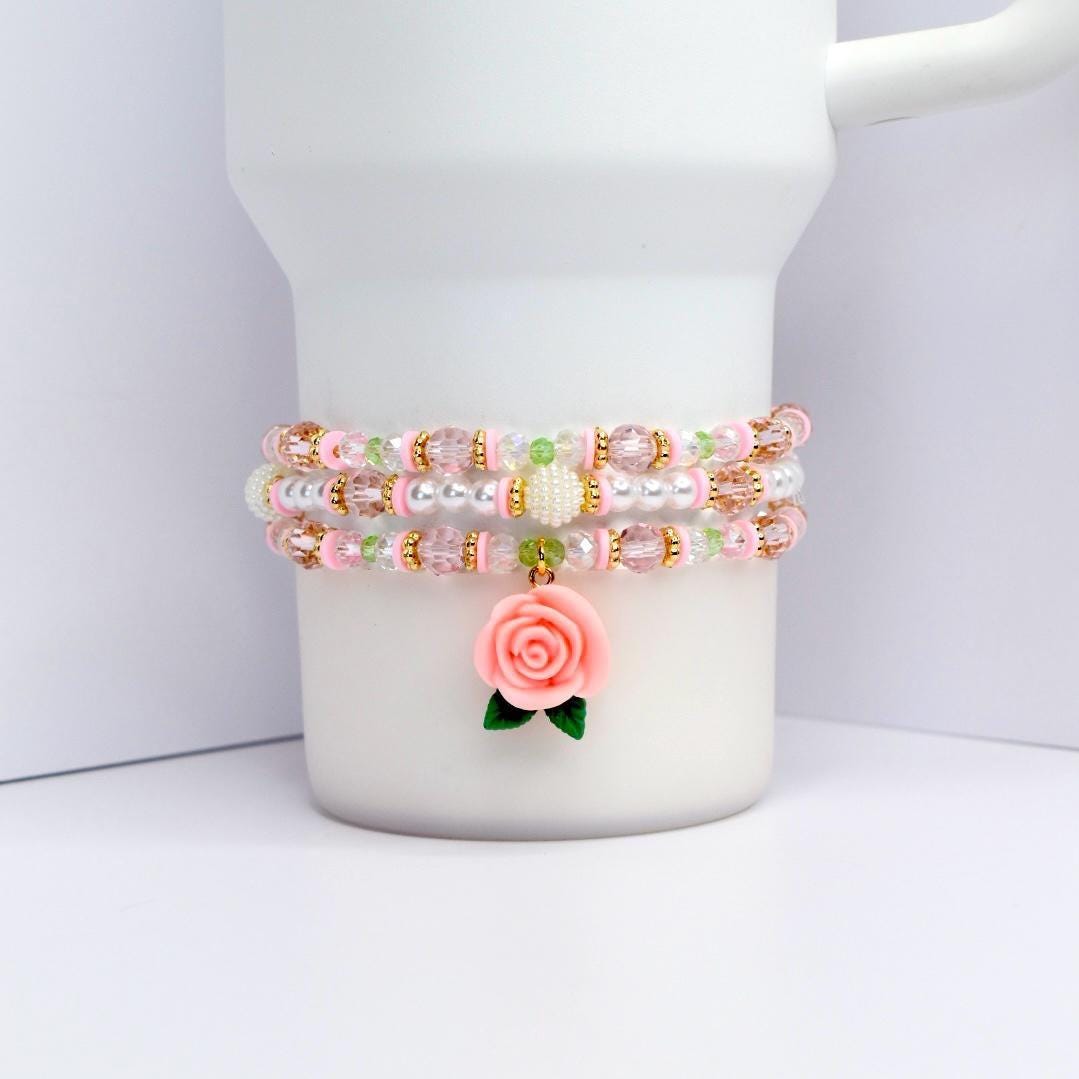 A beaded bracelet set with three strands. The bracelets are pink, white, and gold, with clear and pink beads. A pink rose charm with green leaves hangs from the bottom strand. The bracelets are displayed on a white surface.
