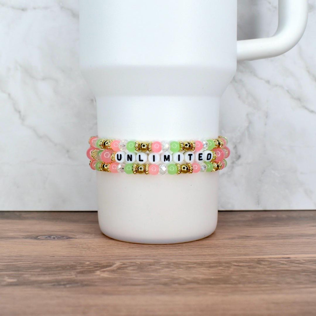 Two beaded bracelets with the word "UNLIMITED" spelled out in white letter beads. The bracelets feature pink, green, and clear beads, with gold-colored accents. The bracelets are displayed on a white tumbler.