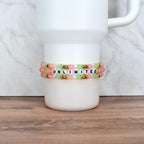 Two beaded bracelets with the word "UNLIMITED" spelled out in white letter beads. The bracelets feature pink, green, and clear beads, with gold-colored accents. The bracelets are displayed on a white tumbler.