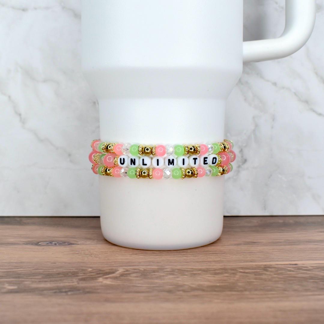 Two beaded bracelets with the word "UNLIMITED" spelled out in white letter beads. The bracelets feature pink, green, and clear beads, with gold-colored accents. The bracelets are displayed on a white tumbler.
