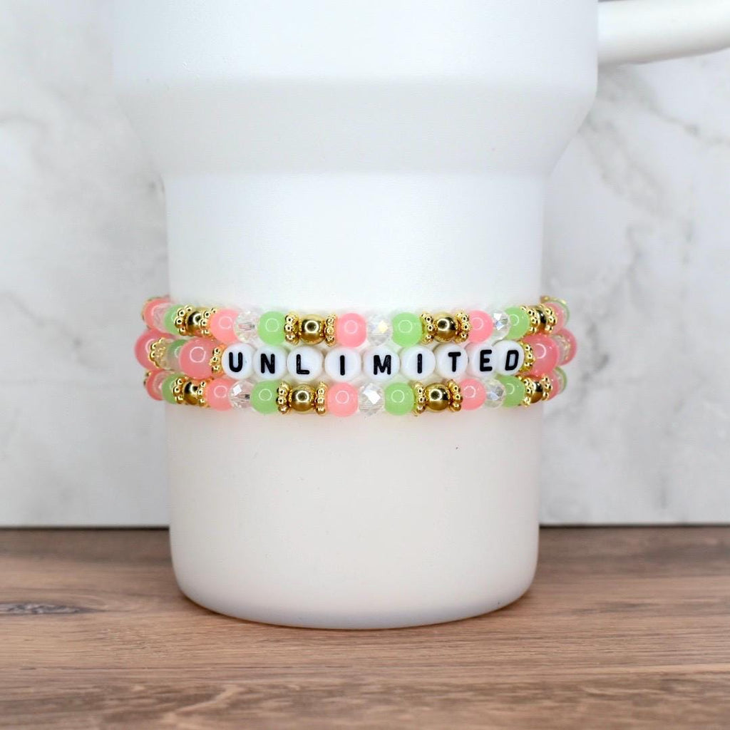 Three beaded bracelets in shades of pink, green, and clear, with gold accents. The center bracelet spells out the word "UNLIMITED" in black and white letter beads. The bracelets are stacked on a white cylindrical object.