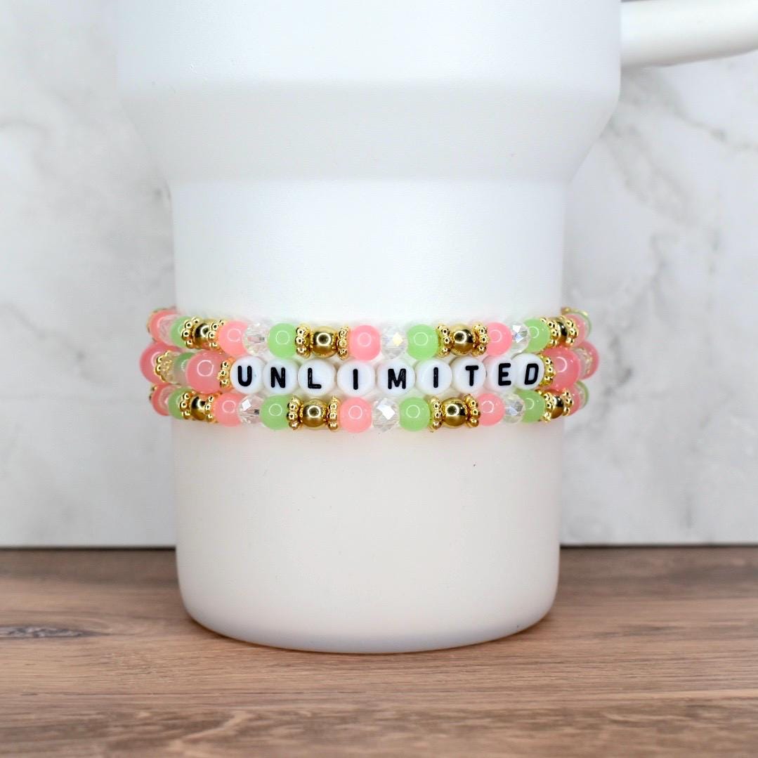 Three beaded bracelets in shades of pink, green, and clear, with gold accents. The center bracelet spells out the word "UNLIMITED" in black and white letter beads. The bracelets are stacked on a white cylindrical object.