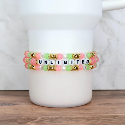 Three beaded bracelets in shades of pink, green, and clear, with gold accents. The center bracelet spells out the word "UNLIMITED" in black and white letter beads. The bracelets are stacked on a white cylindrical object.