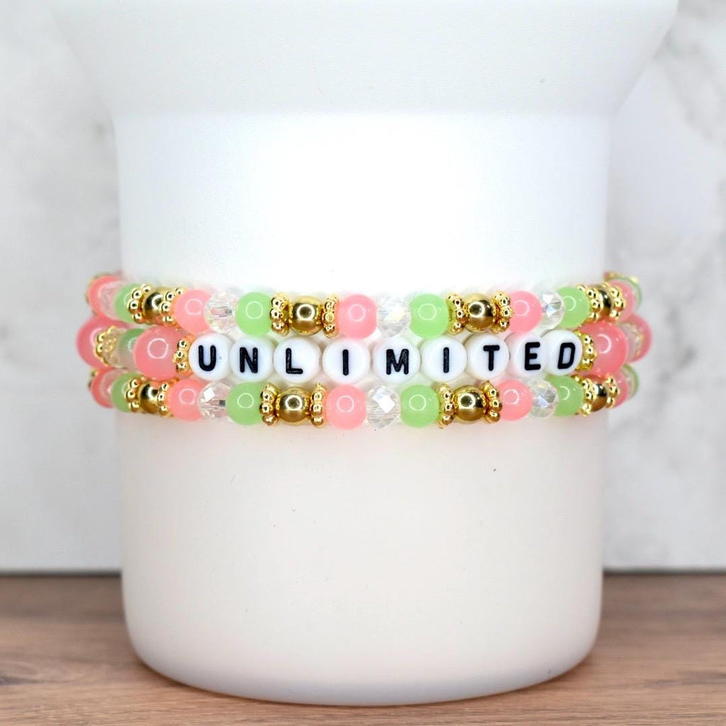 Three beaded bracelets stacked on top of each other. The bracelets feature pink, green, and clear beads, with gold accents. The word "UNLIMITED" is spelled out in black and white letter beads.