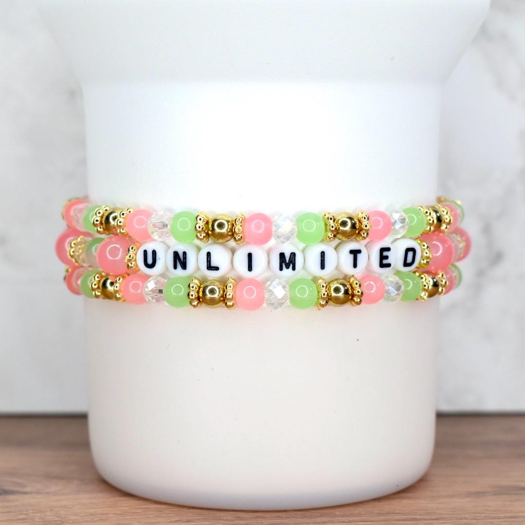 Three beaded bracelets stacked on top of each other. The bracelets feature pink, green, and clear beads, with gold accents. The word "UNLIMITED" is spelled out in black and white letter beads.