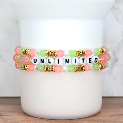 Three beaded bracelets stacked on top of each other. The bracelets feature pink, green, and clear beads, with gold accents. The word "UNLIMITED" is spelled out in black and white letter beads.