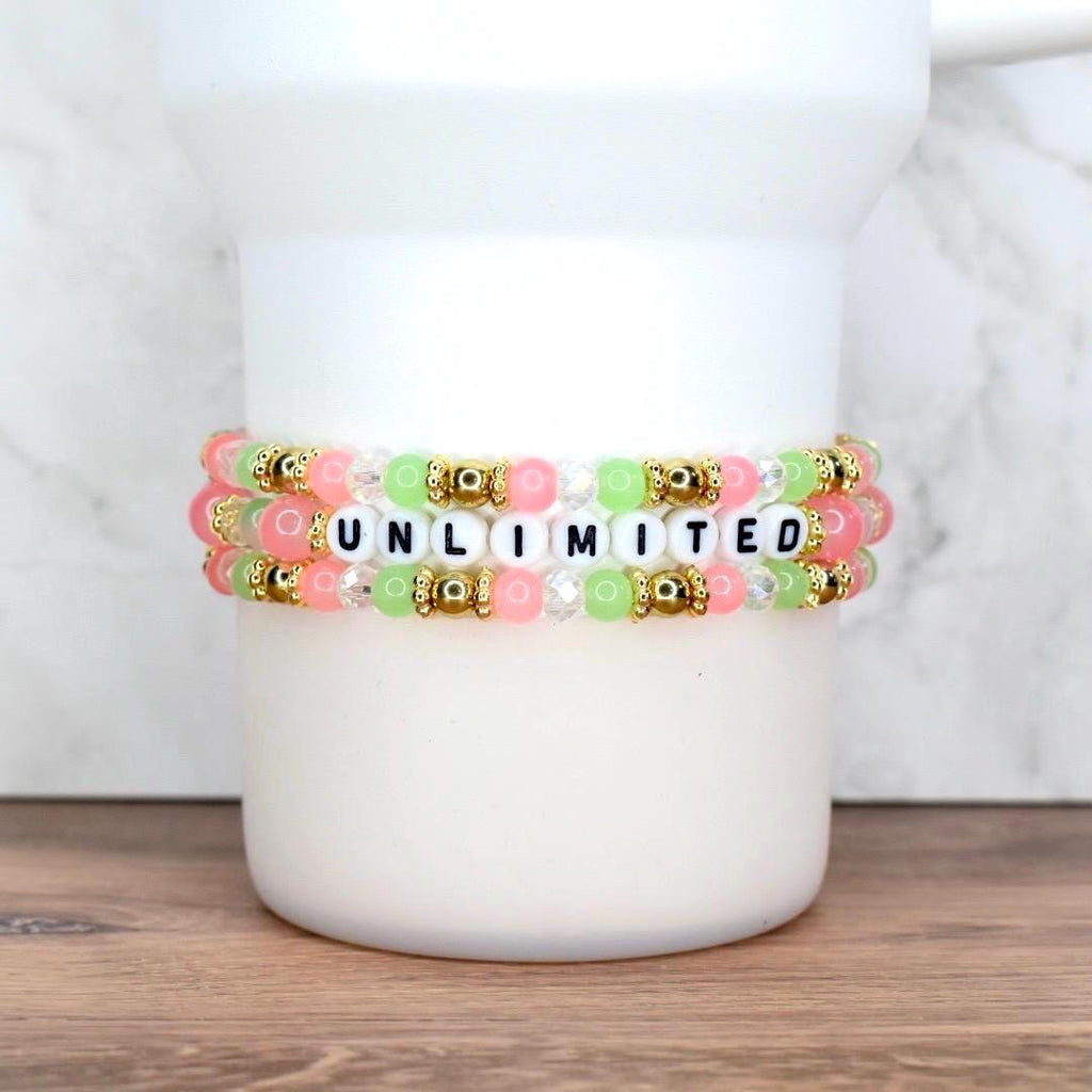 Three beaded bracelets in pink, green, and clear beads with gold accents. The bracelets spell out the word "UNLIMITED" in black letter beads. The bracelets are stacked on a white cylindrical object.