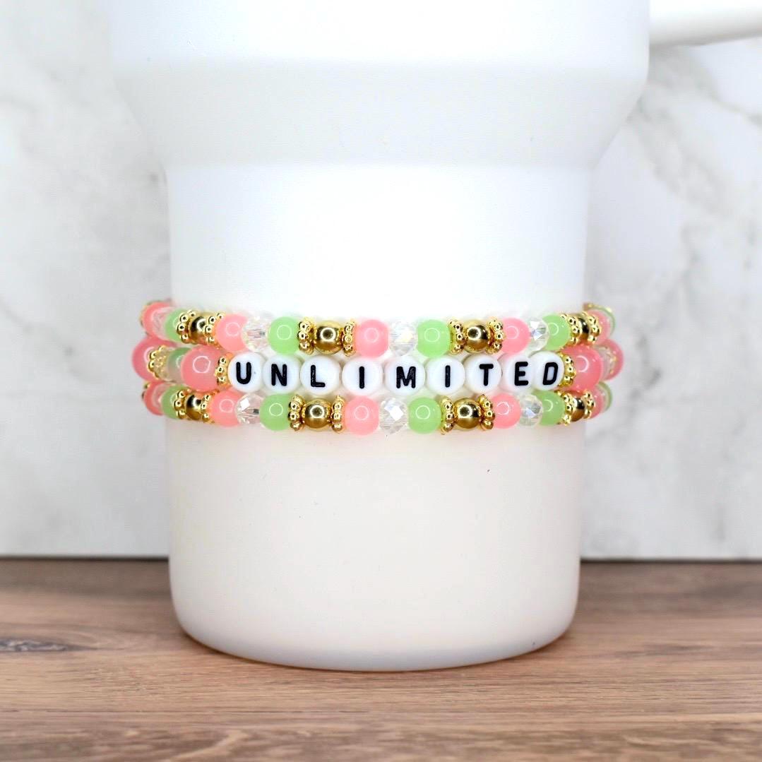 Three beaded bracelets in pink, green, and clear beads with gold accents. The bracelets spell out the word "UNLIMITED" in black letter beads. The bracelets are stacked on a white cylindrical object.