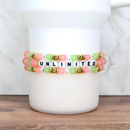 Three beaded bracelets in pink, green, and clear beads with gold accents. The bracelets spell out the word "UNLIMITED" in black letter beads. The bracelets are stacked on a white cylindrical object.