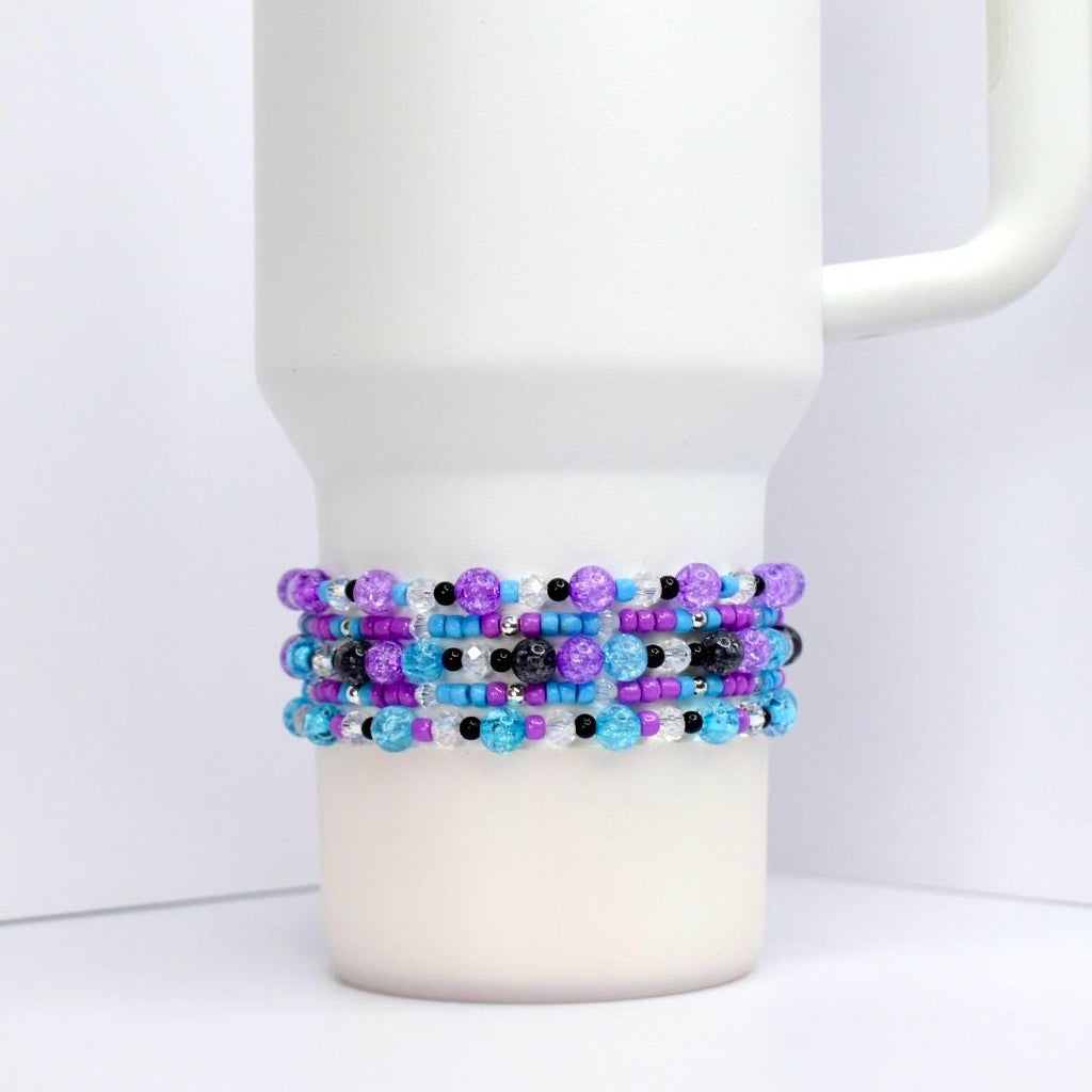 A white tumbler with a beaded wrap. The wrap features rows of purple, blue, black, and clear beads. The beads are arranged in a pattern around the tumbler, creating a decorative accessory.