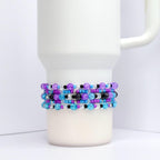A white tumbler with a beaded wrap. The wrap features rows of purple, blue, black, and clear beads. The beads are arranged in a pattern around the tumbler, creating a decorative accessory.