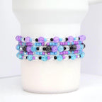 Stack of beaded bracelets in shades of purple, blue, and clear. The bracelets feature a variety of bead sizes and textures, including crackle beads and small black beads. The bracelets are stacked on a white cylindrical object.