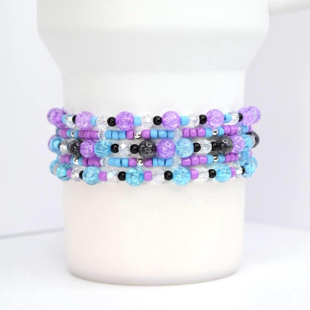 Stack of beaded bracelets in shades of purple, blue, and clear. The bracelets feature a variety of bead sizes and textures, including crackle beads and small black beads. The bracelets are stacked on a white cylindrical object.