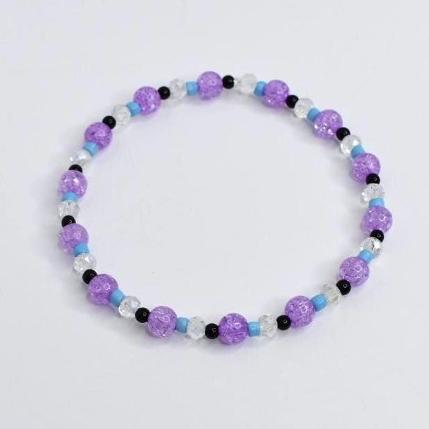 A beaded bracelet featuring a mix of purple, clear, black, and light blue beads. The bracelet is made with crackle-style purple beads, interspersed with clear, black, and light blue beads. This stretch bracelet is a piece of jewelry.