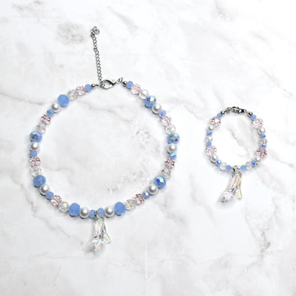 A necklace and bracelet set featuring light blue, pink, and clear beads. Each piece has a silver-toned chain and a charm shaped like a glass slipper. The jewelry is displayed on a white marble surface.