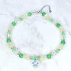 A beaded necklace featuring green, yellow, and clear beads. The necklace has a silver chain and a heart-shaped crystal pendant. The beads are various sizes and the necklace is displayed on a white marble surface.