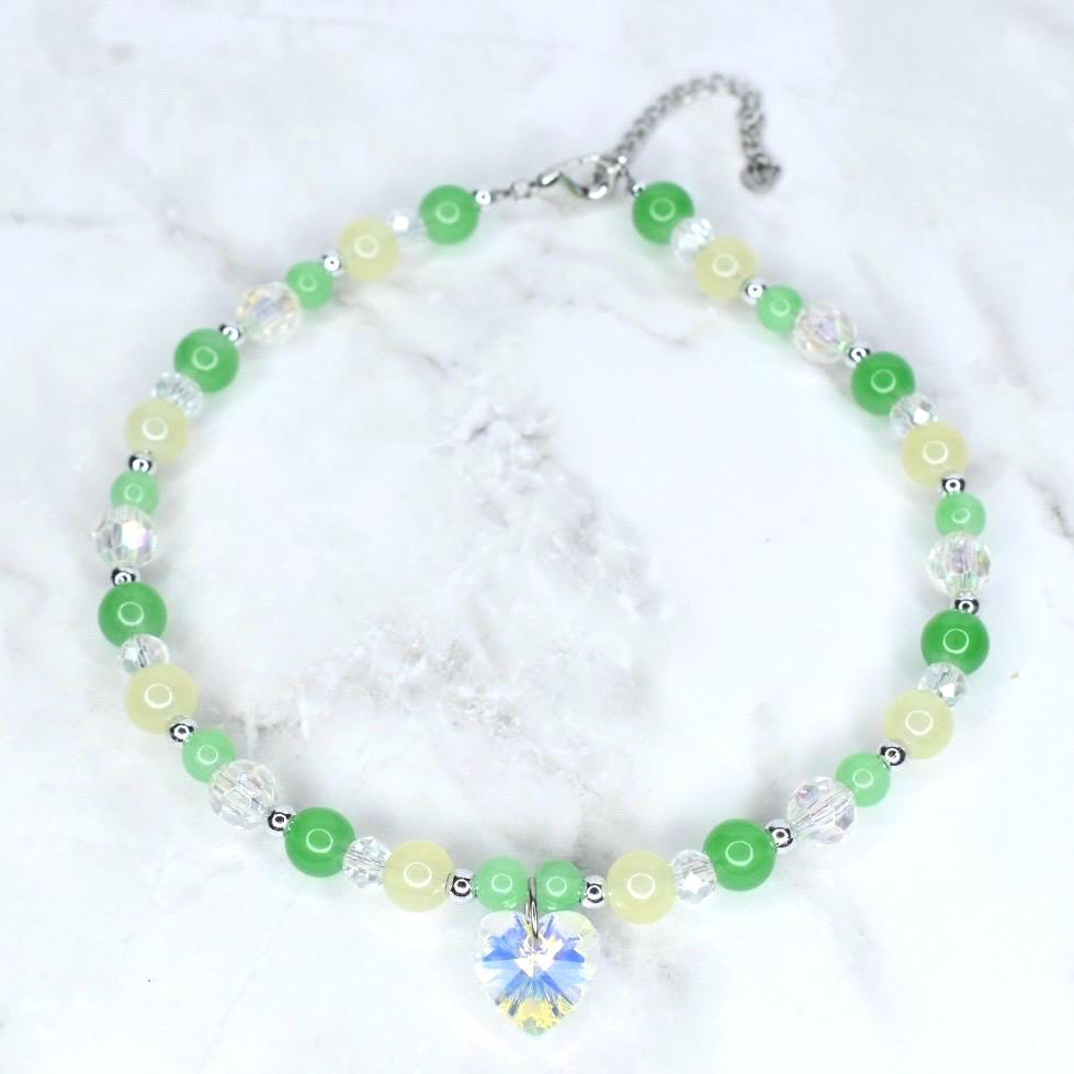 A beaded necklace featuring green, yellow, and clear beads. The necklace has a silver chain and a heart-shaped crystal pendant. The beads are various sizes and the necklace is displayed on a white marble surface.