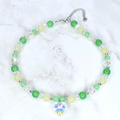 A beaded necklace featuring green, yellow, and clear beads. The necklace has a silver chain and a heart-shaped crystal pendant. The beads are various sizes and the necklace is displayed on a white marble surface.
