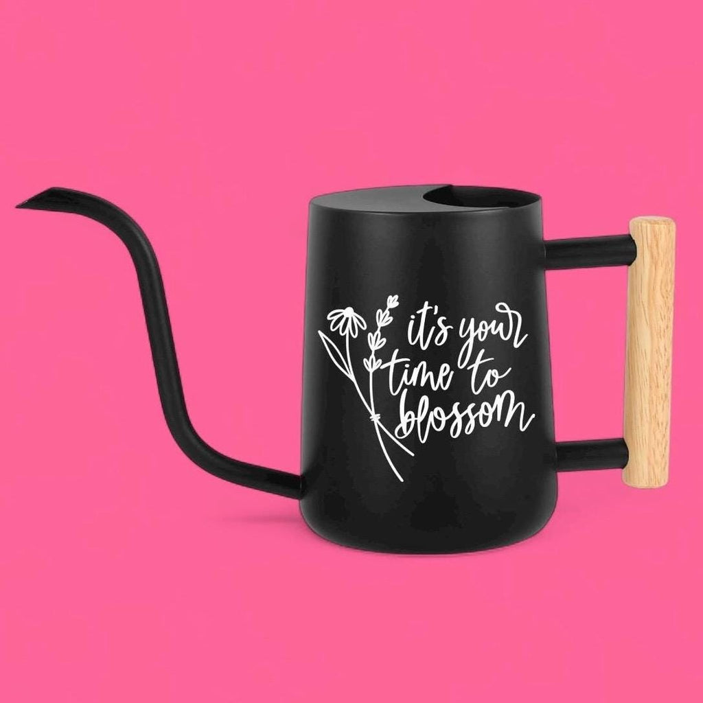 Black watering can with a long, curved spout and a wooden handle. The can features white text that reads 'it's your time to blossom' with a floral design. The handle is made of light-colored wood, and the can has a sleek, modern design, suitable for indoor or outdoor gardening.
