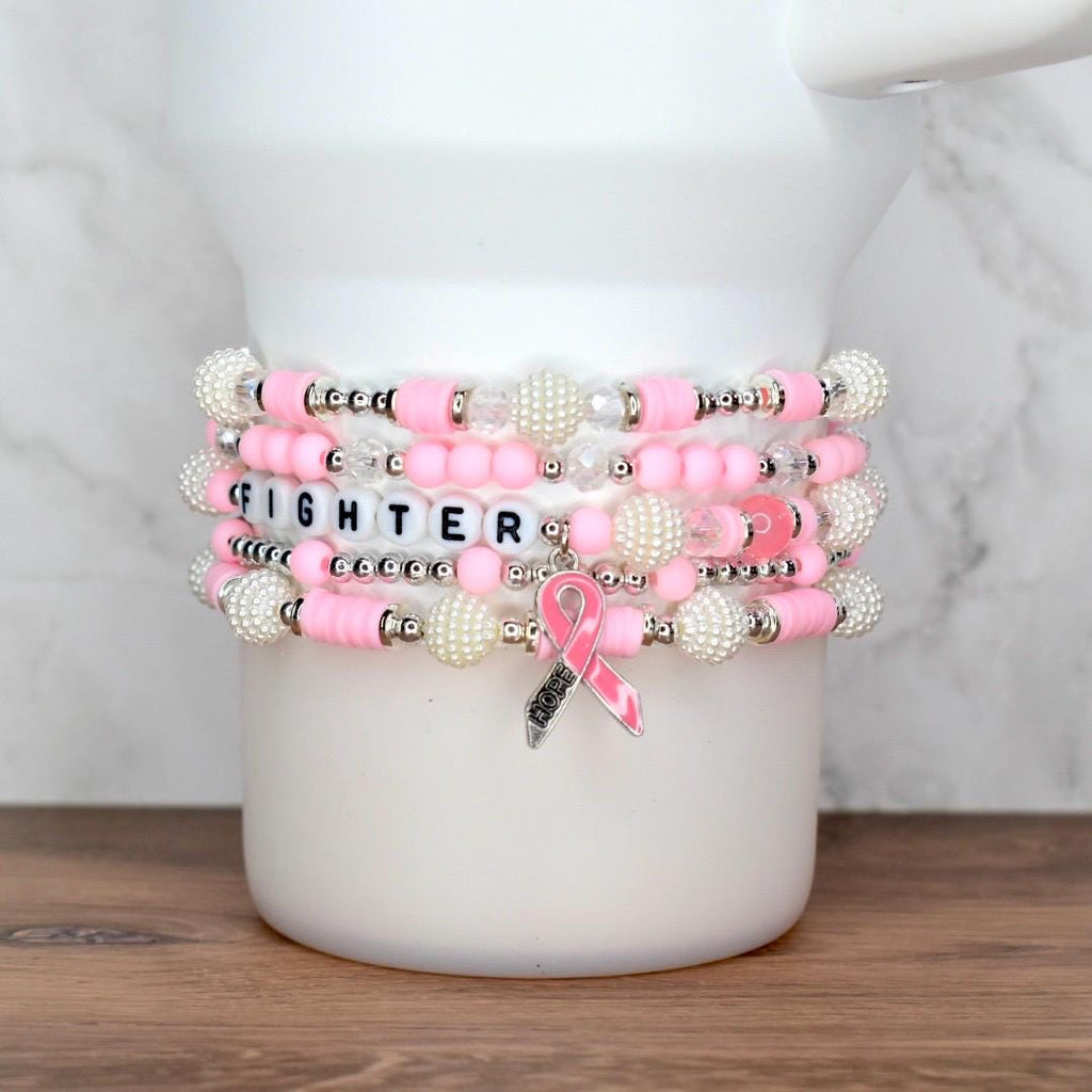 Stack of pink and white beaded bracelets. One bracelet spells "FIGHTER" with letter beads. A pink ribbon charm with the word "HOPE" hangs from one bracelet. The bracelets are stacked on a white surface.