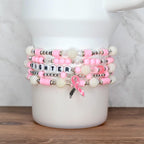 Stack of pink and white beaded bracelets. One bracelet spells "FIGHTER" with letter beads. A pink ribbon charm with the word "HOPE" hangs from one bracelet. The bracelets are stacked on a white surface.