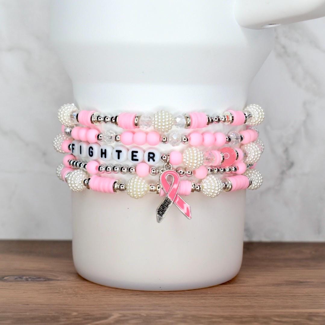 Stack of pink and white beaded bracelets. One bracelet spells "FIGHTER" with letter beads. A pink ribbon charm with the word "HOPE" hangs from one bracelet. The bracelets are stacked on a white surface.