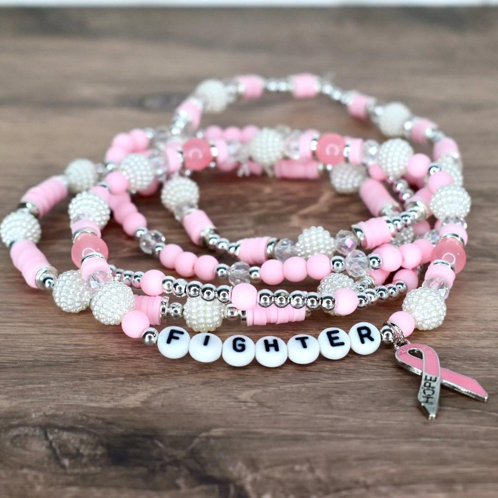 Pink beaded bracelets with white pearl accents and silver beads. One bracelet spells out "FIGHTER" in white letter beads. A pink ribbon charm with the word "HOPE" hangs from the bracelet.