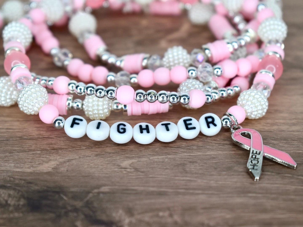 Pink and white beaded bracelets with the word "FIGHTER" spelled out in white letter beads. A pink ribbon charm with the word "HOPE" hangs from the bracelet. The bracelets are on a wooden surface.