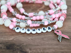 Pink and white beaded bracelets with the word "FIGHTER" spelled out in white letter beads. A pink ribbon charm with the word "HOPE" hangs from the bracelet. The bracelets are on a wooden surface.