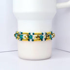 A beaded bracelet with gold, teal, and clear beads. The bracelet is wrapped around a white mug. The beads are arranged in rows, with gold beads on the top and bottom rows and teal and clear beads in the middle.