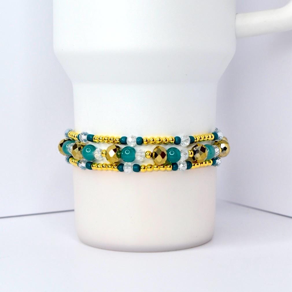 A beaded bracelet with gold, teal, and clear beads. The bracelet is wrapped around a white mug. The beads are arranged in rows, with gold beads on the top and bottom rows and teal and clear beads in the middle.