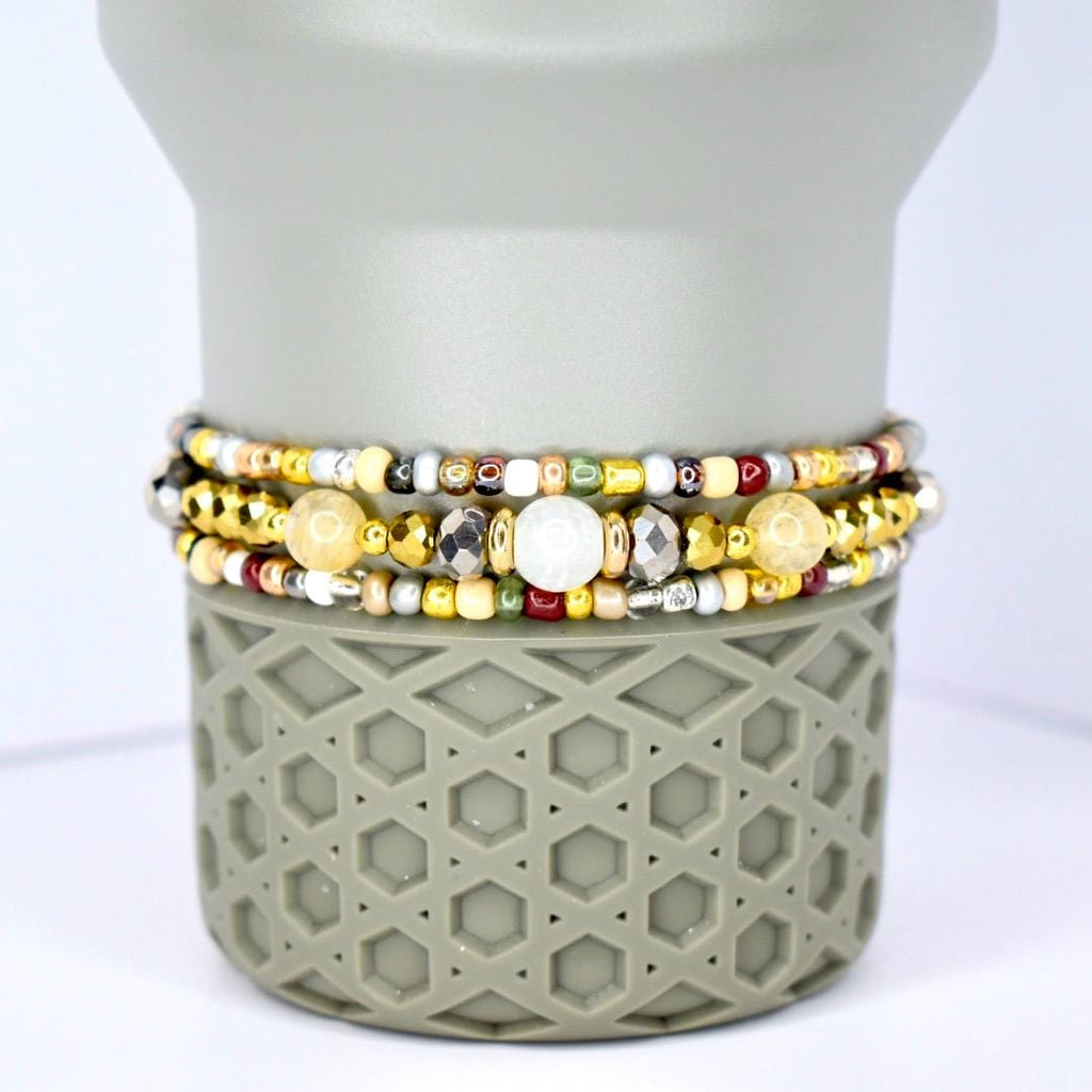 Three beaded bracelets in gold, silver, and cream colors. The bracelets feature a variety of beads, including small gold, silver, and cream-colored beads, and larger, faceted beads. The bracelets are stacked on a gray, geometric-patterned base.