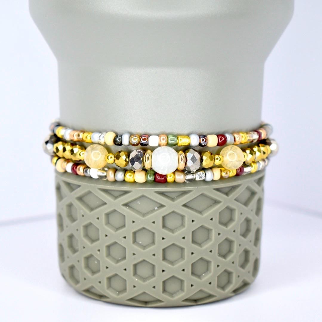 Three beaded bracelets in gold, silver, and cream colors. The bracelets feature a variety of beads, including small gold, silver, and cream-colored beads, and larger, faceted beads. The bracelets are stacked on a gray, geometric-patterned base.