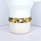 Three beaded bracelets in shades of gold, silver, and cream are stacked on a white cylindrical object. The bracelets feature a variety of small beads, including metallic, opaque, and translucent elements. The bracelets are likely handmade and suitable for everyday wear.